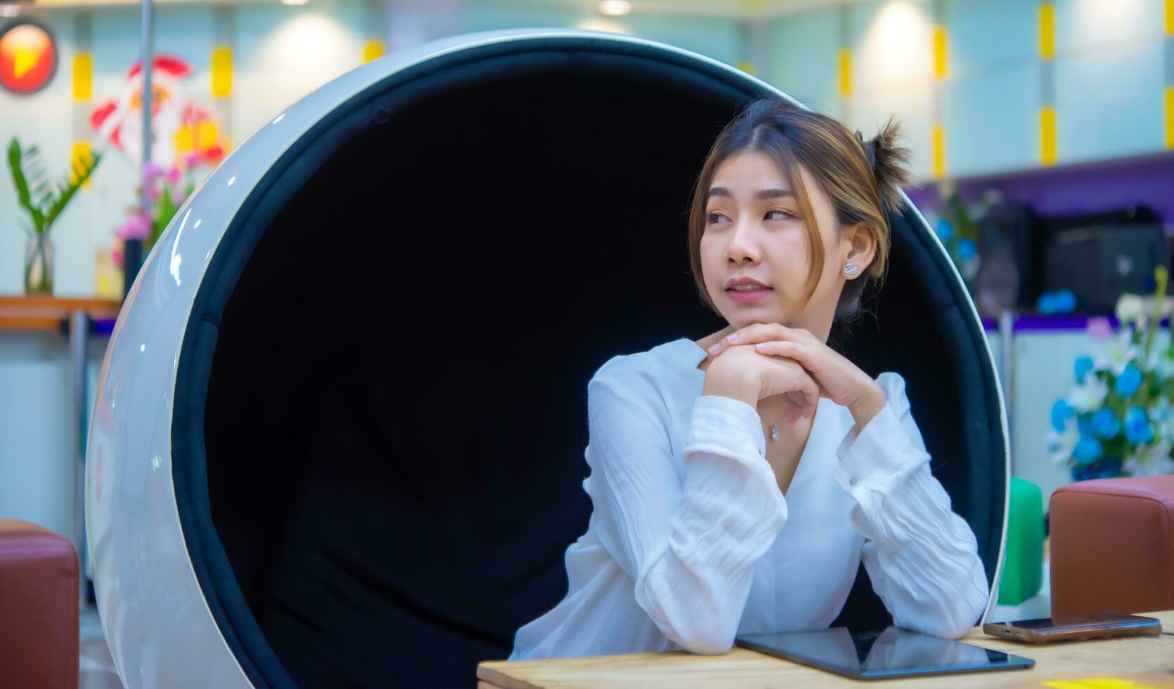 Beautiful asian business woman sitting sideways on a modern round chair appears to be in thought on the table has her phone and tablet and smiling while looking to the copy space, Digital marketing photo