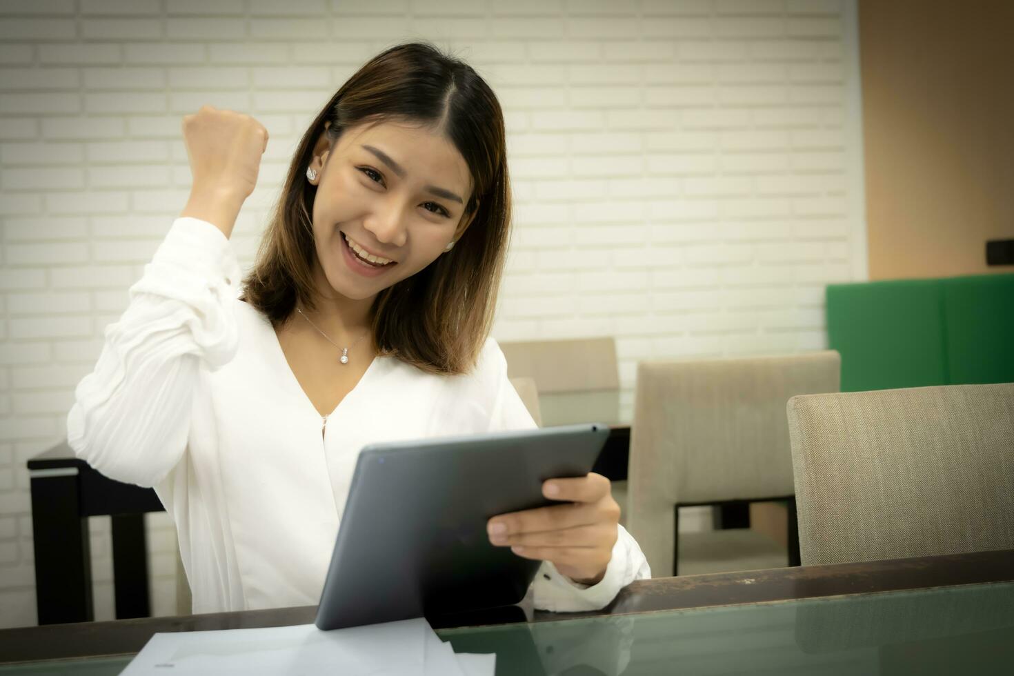 Beautiful asian female teacher is sitting showing celebrate clenching fist while holding tablet with smile on her face. photo
