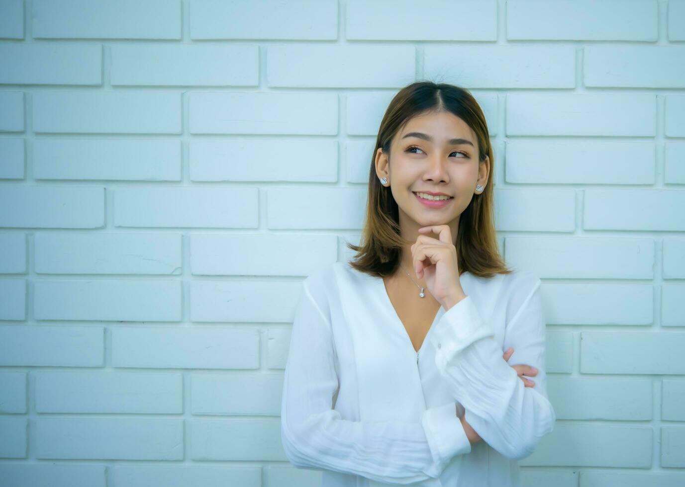 Beautiful asian business woman is standing in a thoughtful pose with a smile at the copy space with a white brick background, Digital marketing. photo