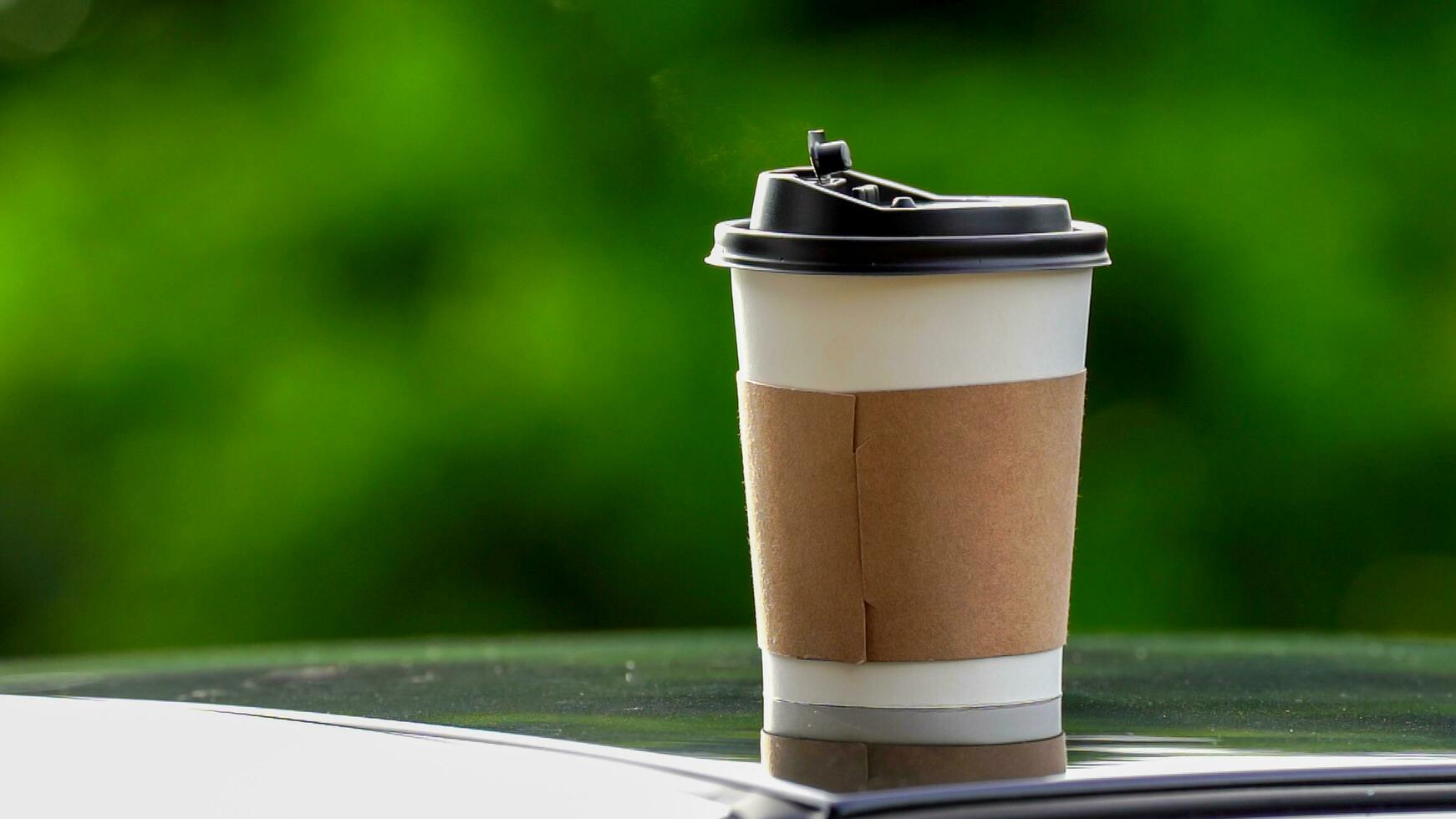 coffee takeaway in a paper cup on top of the car roof green tree background at sunrise in the morning,  selective focus, soft focus. photo