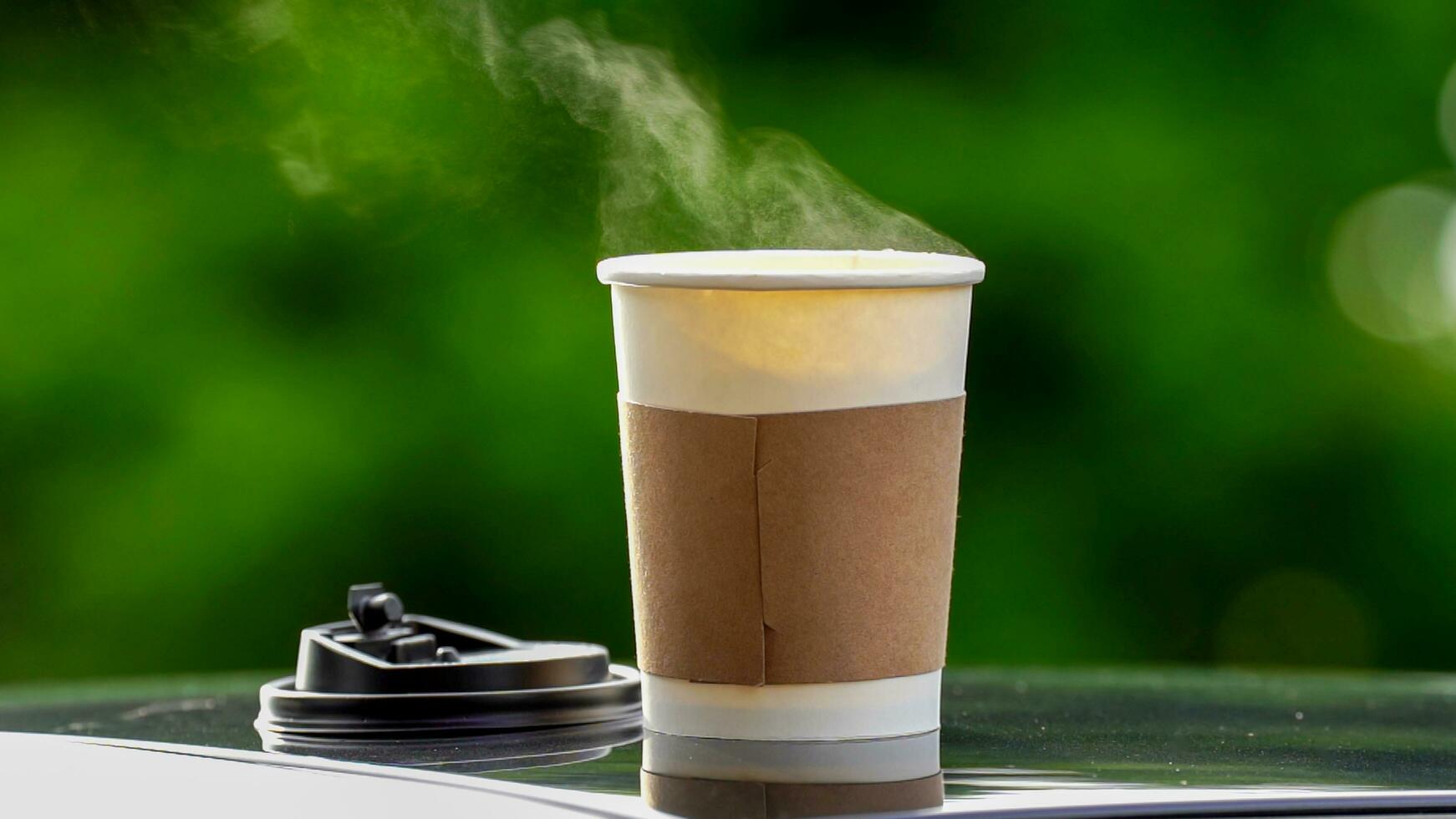 coffee takeaway in a paper cup on top of the car roof green tree background at sunrise in the morning,  selective focus, soft focus. photo