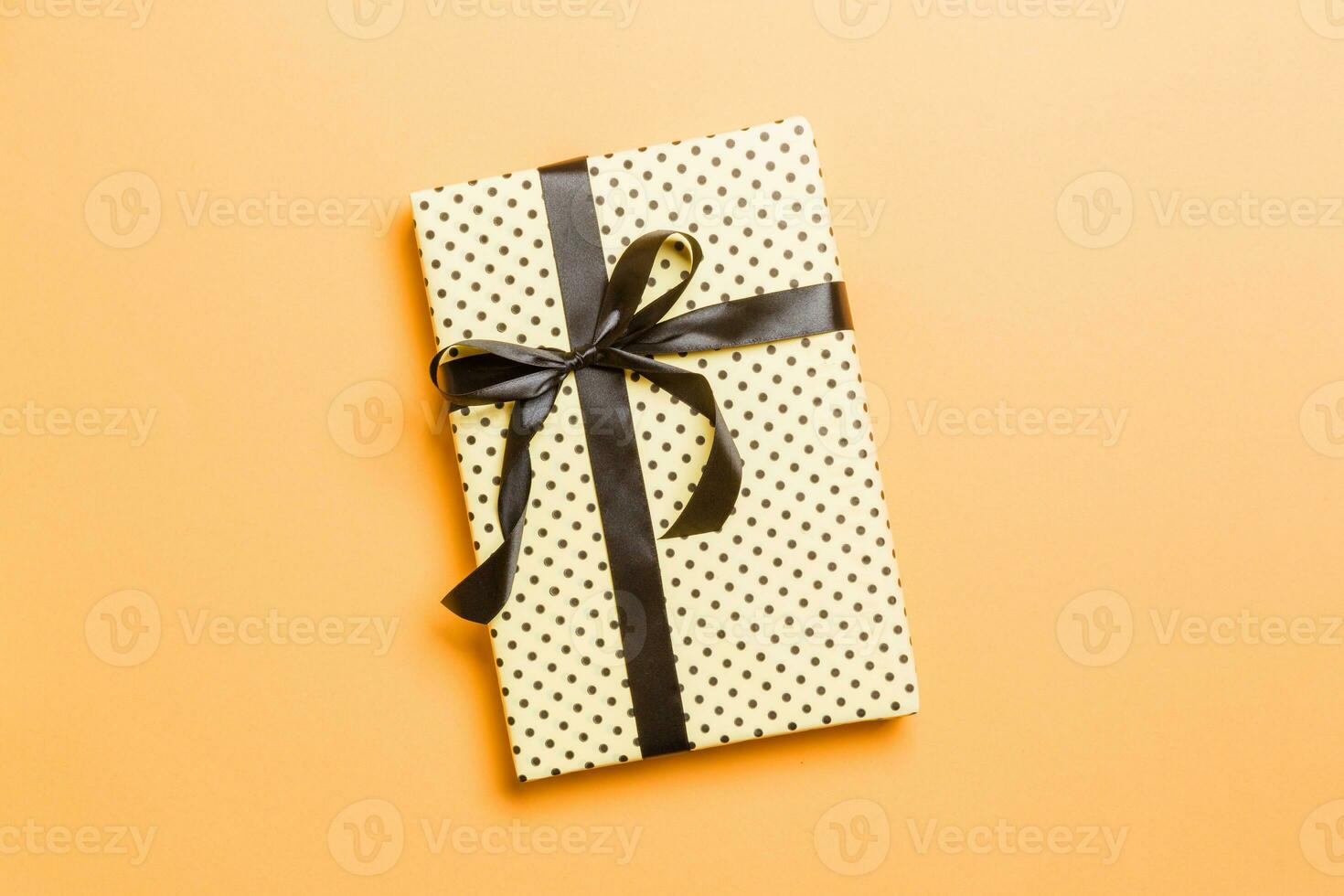 wrapped Christmas or other holiday handmade present in paper with black ribbon on orange background. Present box, decoration of gift on colored table, top view with copy space photo