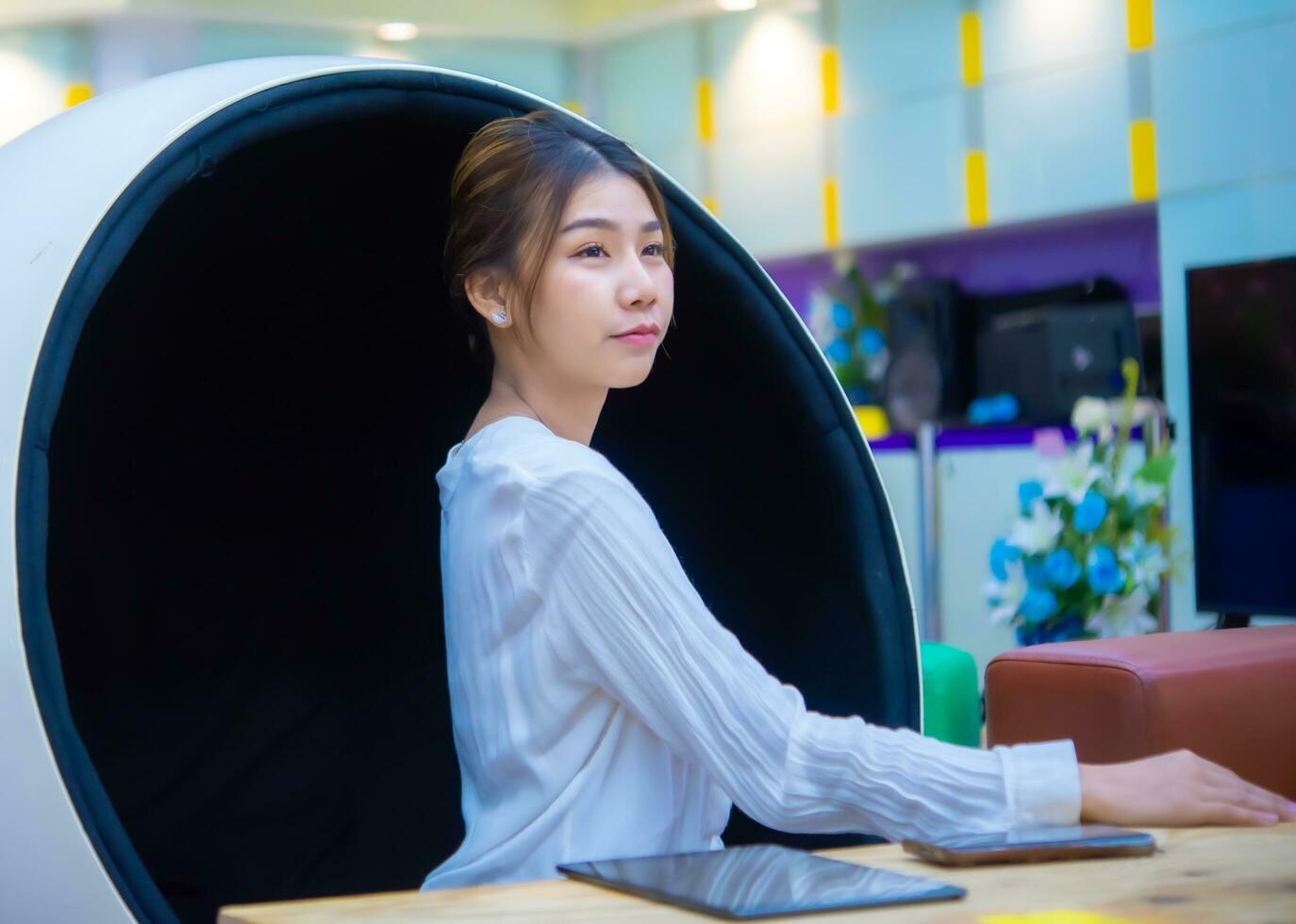 hermosa asiático negocio mujer es sentado en un moderno redondo silla conmovedor el mesa y tiene un teléfono y tableta en eso mientras mirando a el Copiar espacio con calma rostro, digital marketing. foto