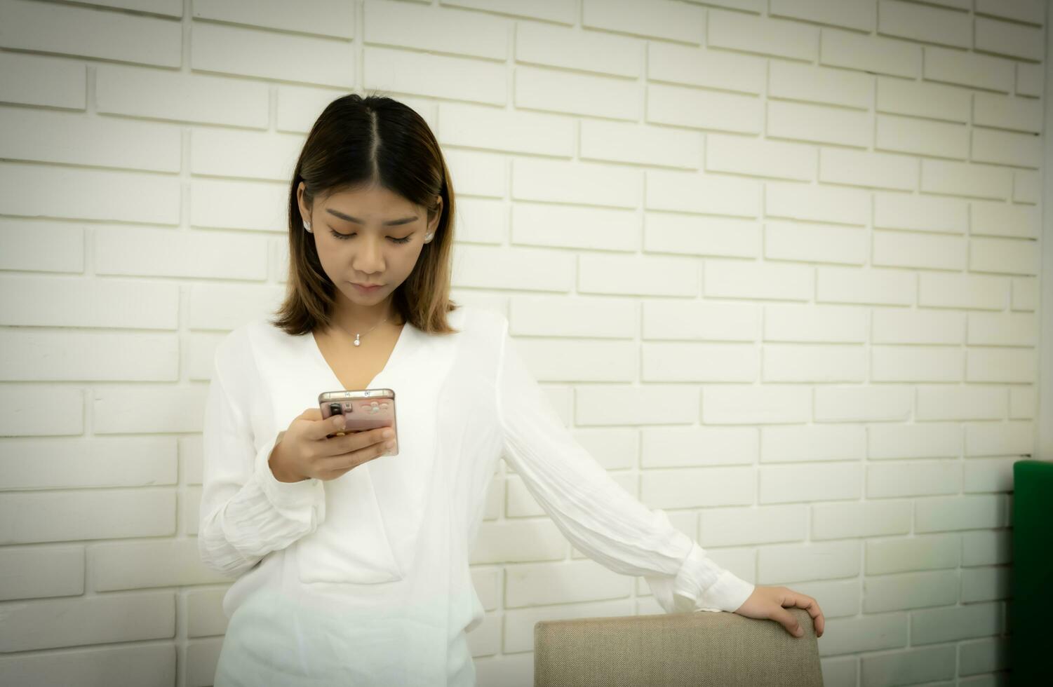 hermosa asiático en línea vendedor es en pie y participación su teléfono a cheque fuera su en línea Tienda con grave cara en contra un blanco ladrillo pared antecedentes. foto