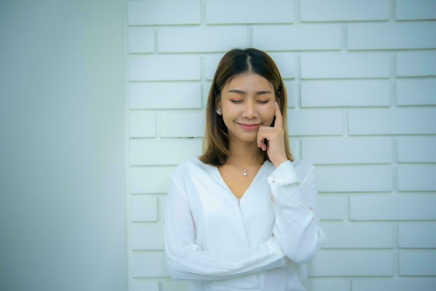 hermosa asiático negocio mujer es en pie en contra un blanco ladrillo muro, con su manos en su las mejillas sonriente y clausura su ojos, digital marketing. foto