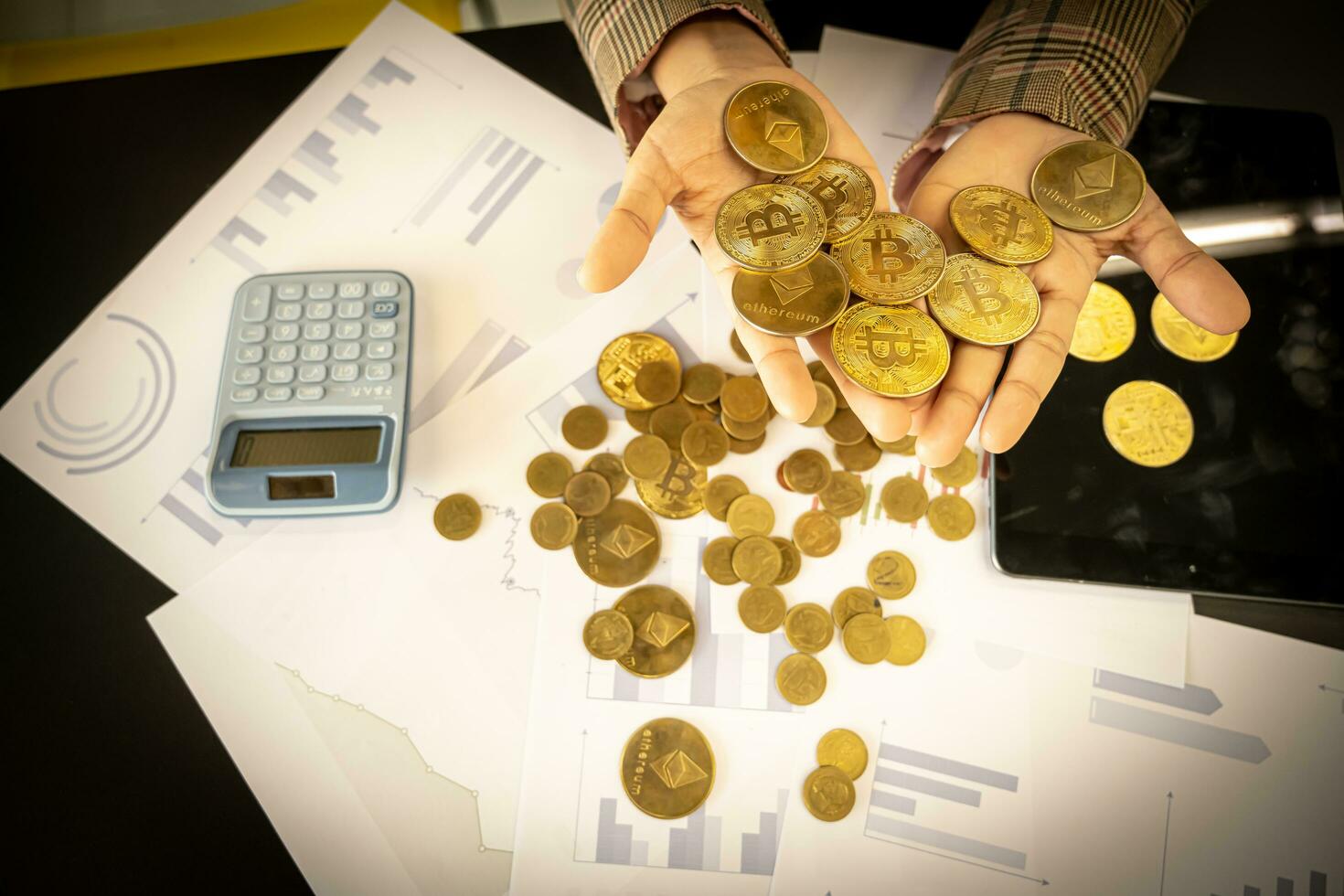 Close-up image of a woman holding golden-colored bitcoins and showing her document with various graphs on her desk. photo