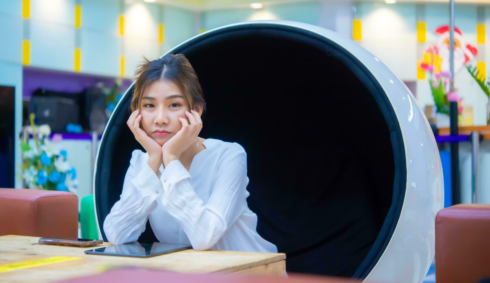 Beautiful asian business woman is sitting on a modern round chair with sad face on the table has a phone and tablet on it while looking at the camera, Digital marketing. photo