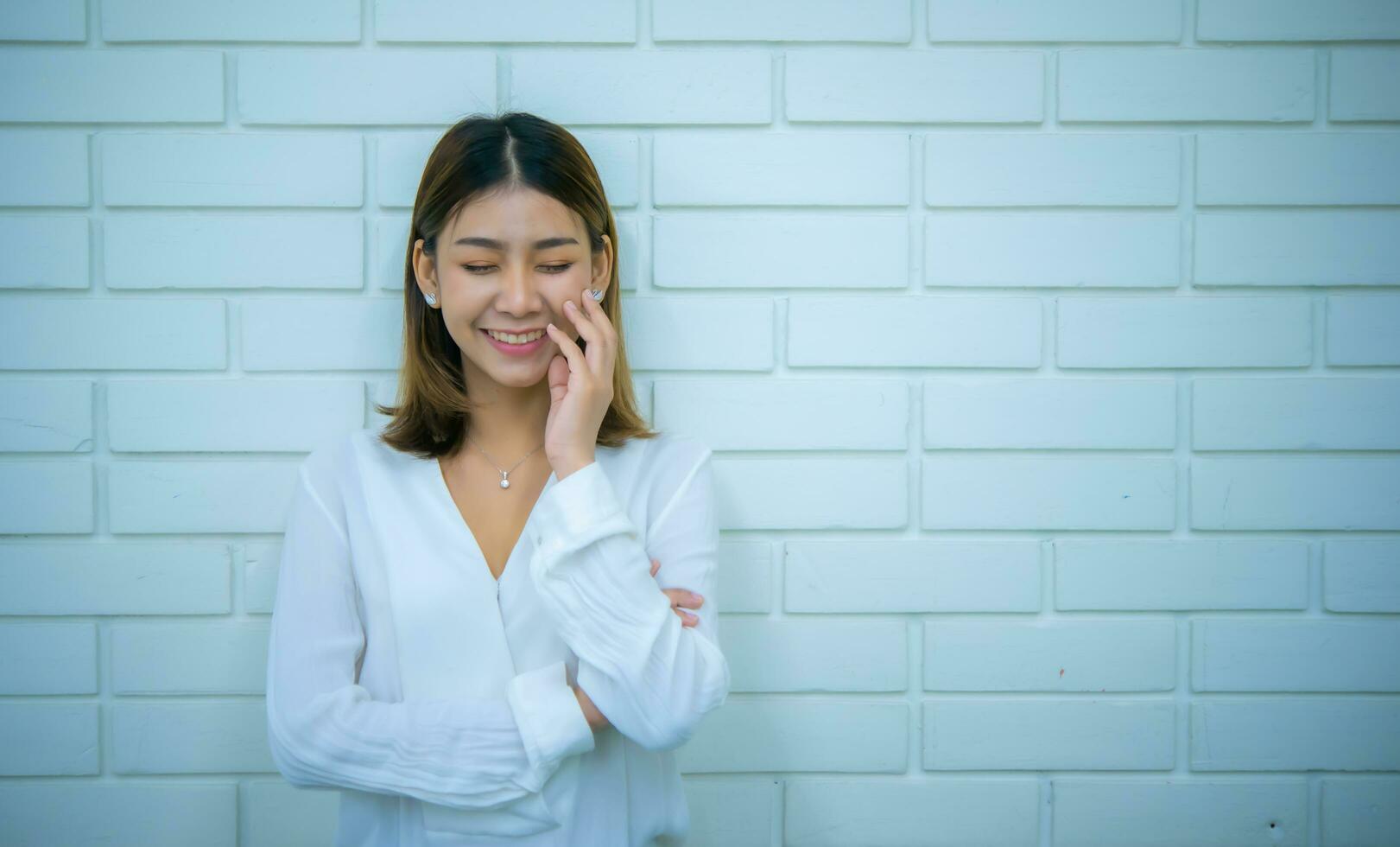 Beautiful asian business woman is standing with one hand on her face smiling while closing her eyes with white bricks in the background, Digital marketing. photo
