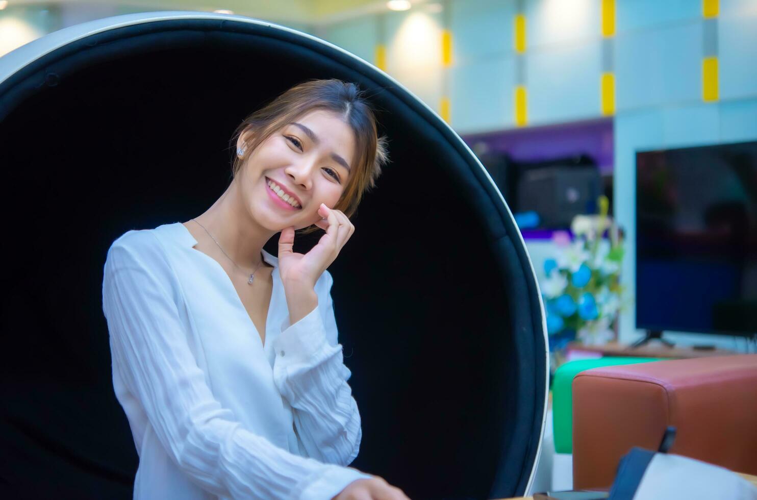 Beautiful asian business woman sitting in a modern round chair with one hand on her cheek and smiling face while looking to the camera, Digital marketing. photo
