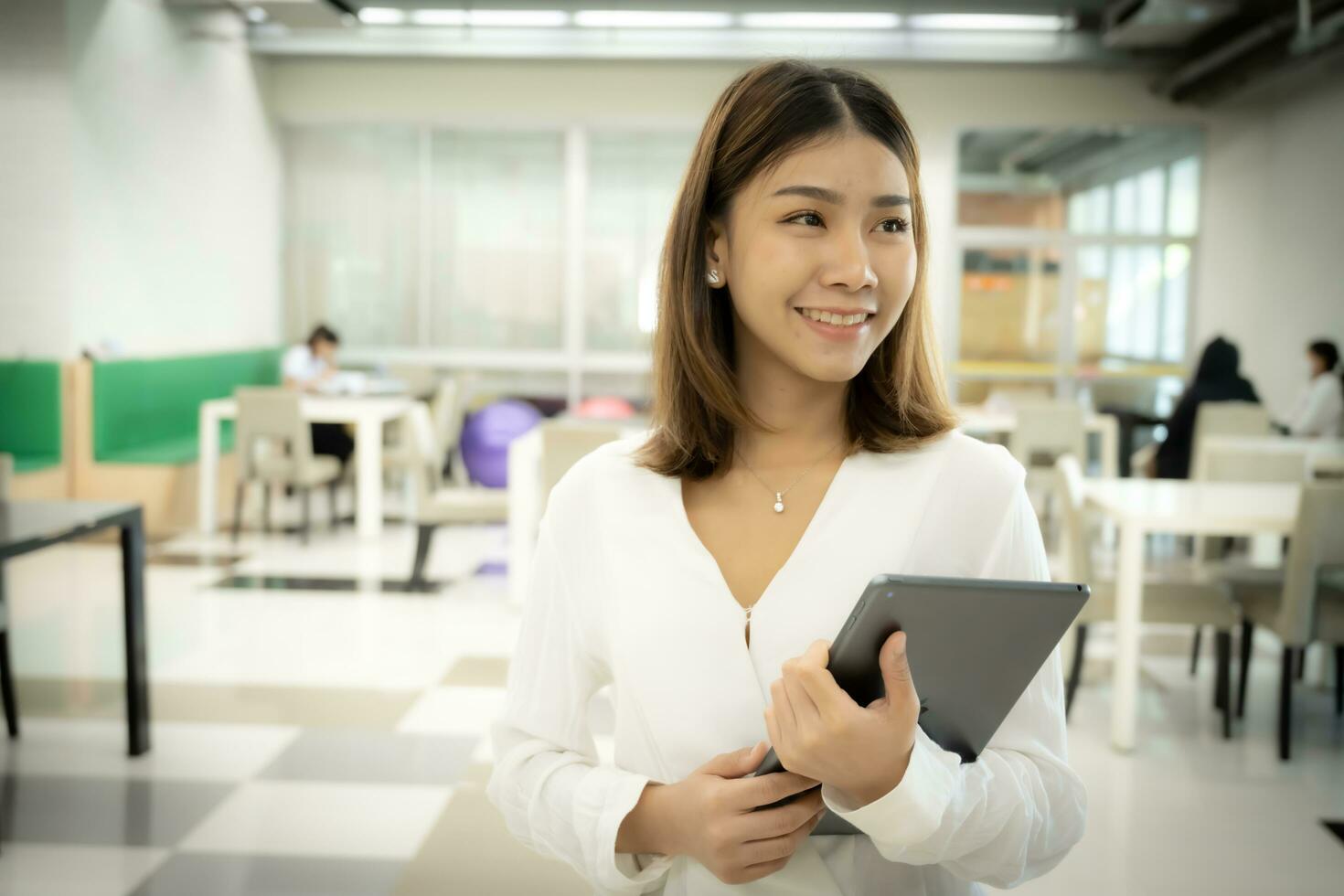 Beautiful asian female teacher is standing while holding tablet with a smiling face in the library room. photo