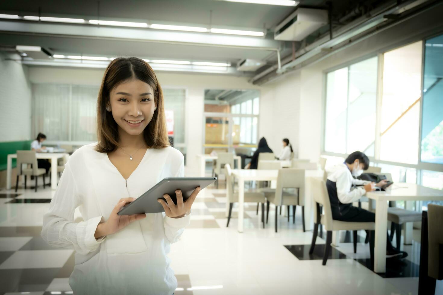Beautiful asian female teacher is looking to the camera while holding tablet with a smiling face in the library room. photo