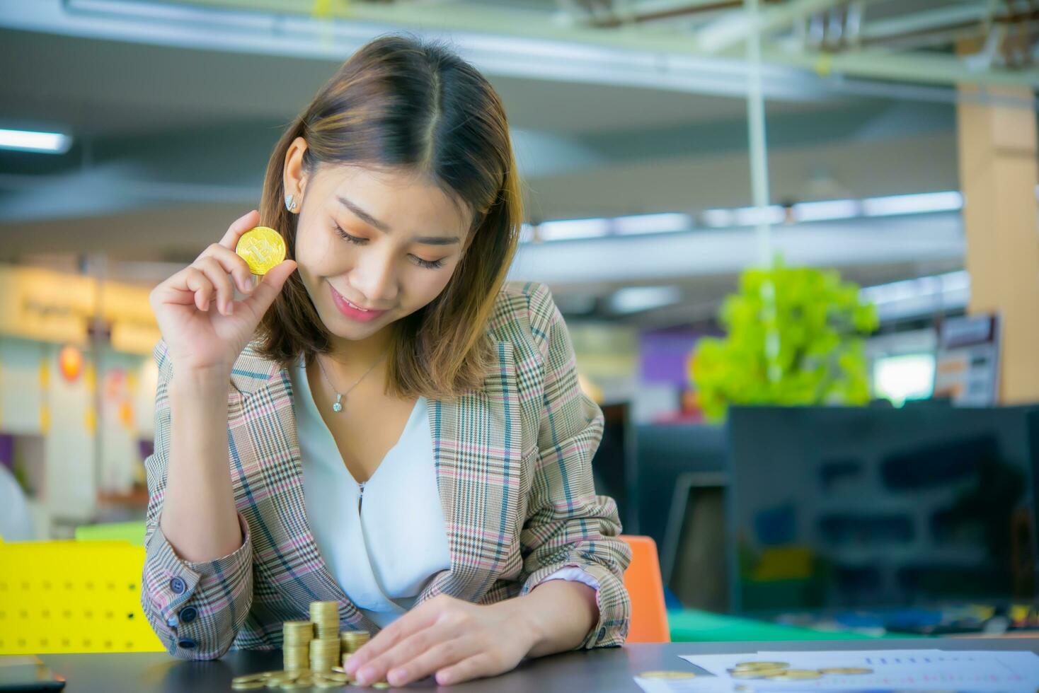 Young beautiful asian business woman holding a coin in one hand while looking at the pile of gold coins with a smile it means that there is good financial management. photo