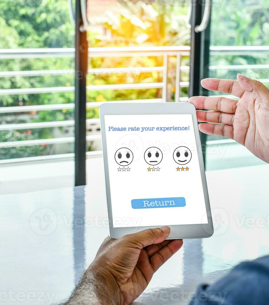 Tablet screen displays a Please rate your experience message to confirm Service satisfaction. A man sits and holding tablet in the work at home. Concept of business service survey. Blurred background photo