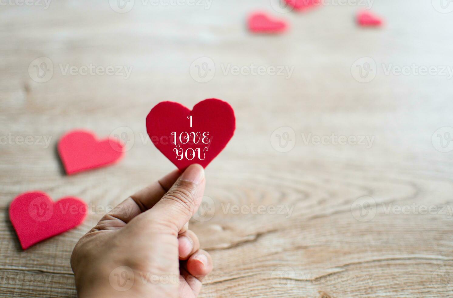 Concept Valentine's day, man's hand holding a red heart symbol, letters I love you white, and red heart symbol blur placed on a wooden table. Close-up, selective focus, copy space for design, and text photo