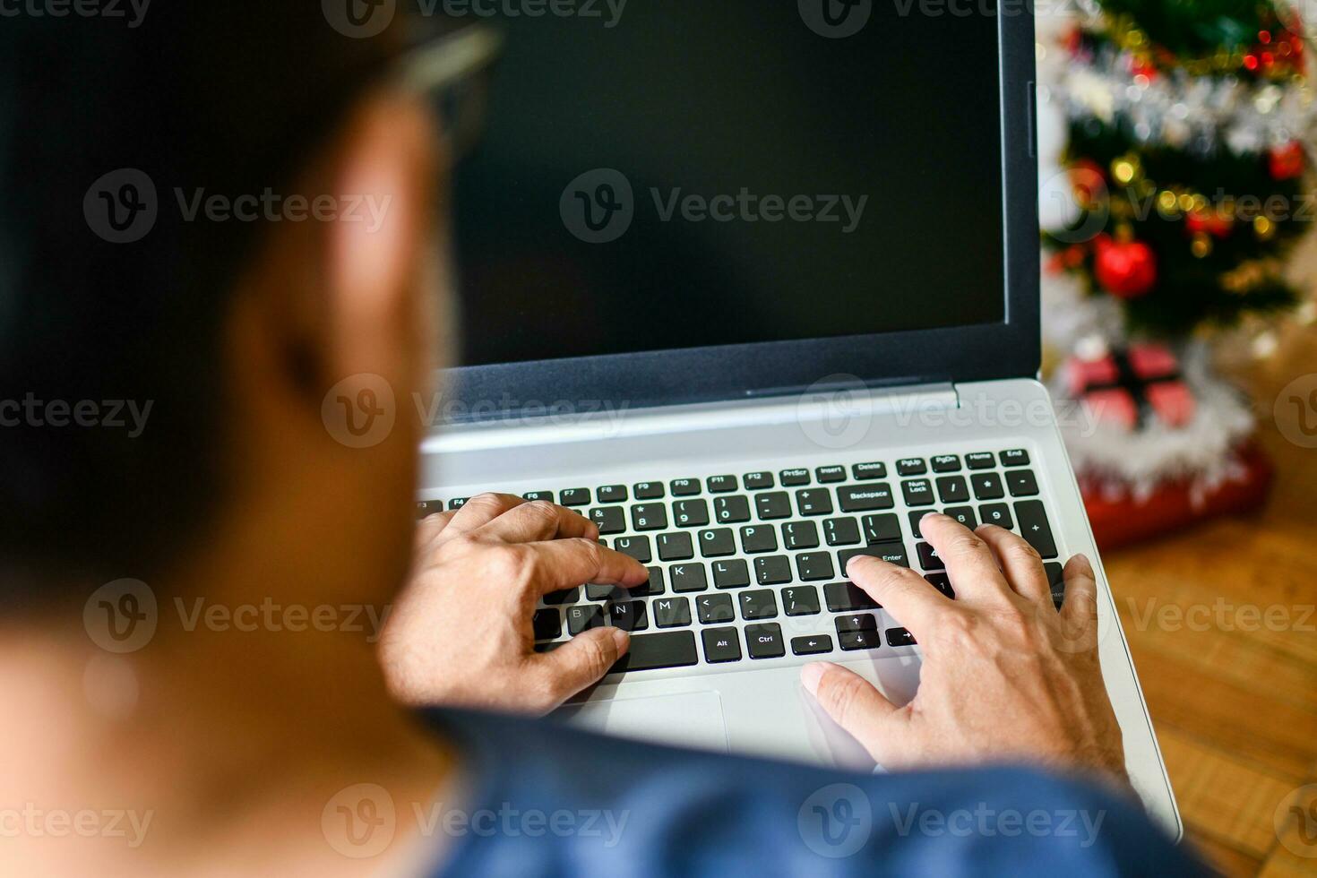 Closeup, People using laptop computer devices at home. A man typing a keyboard notebook computer in the living room at home, Blank black screen for design, Text, Blurred background, and Christmas tree photo