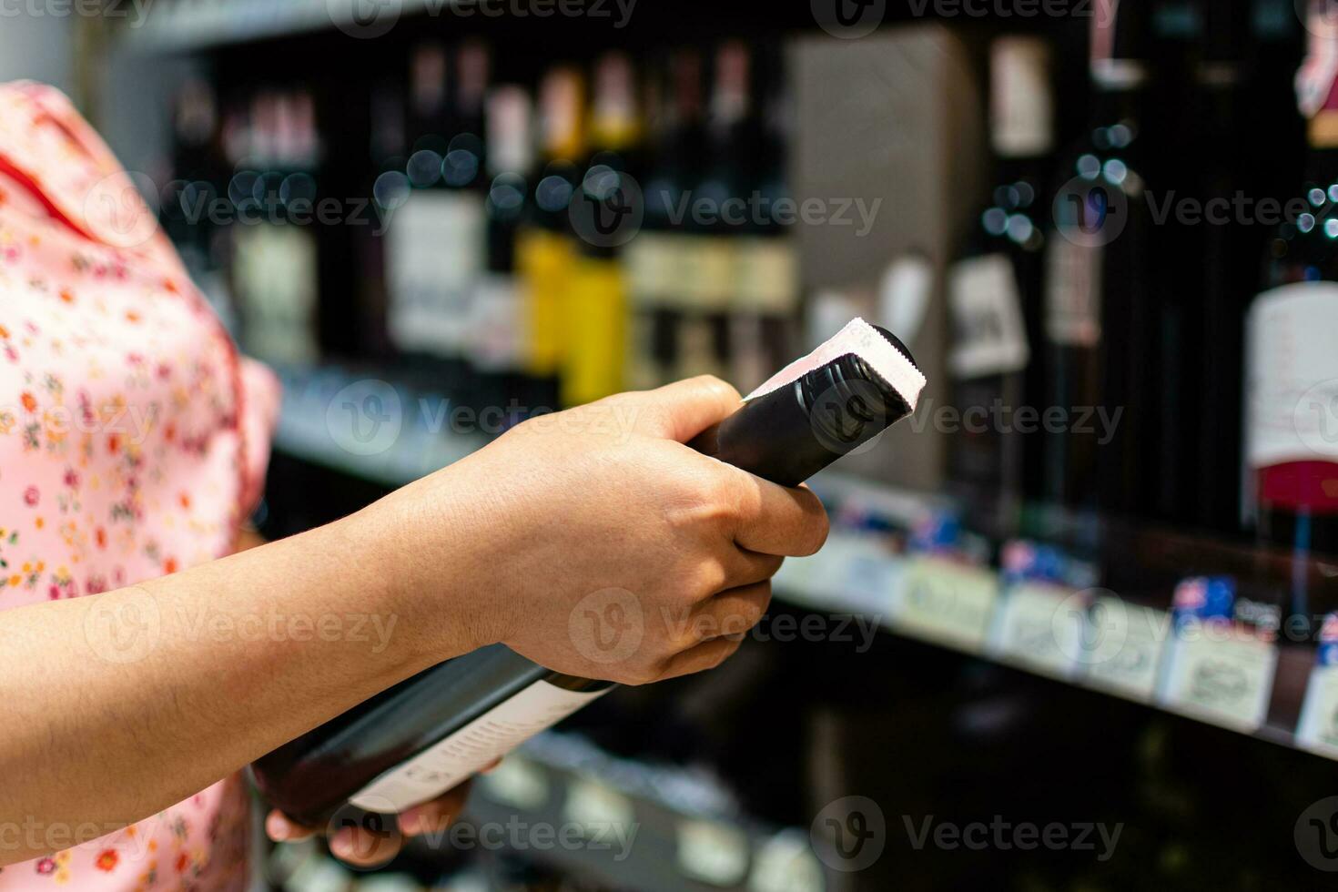 female customer holding, choosing, reading a red wine bottle label in a department store. An Asian woman holds a bottle of wine at the supermarket alcohol section. A shelf full of alcoholic beverages photo