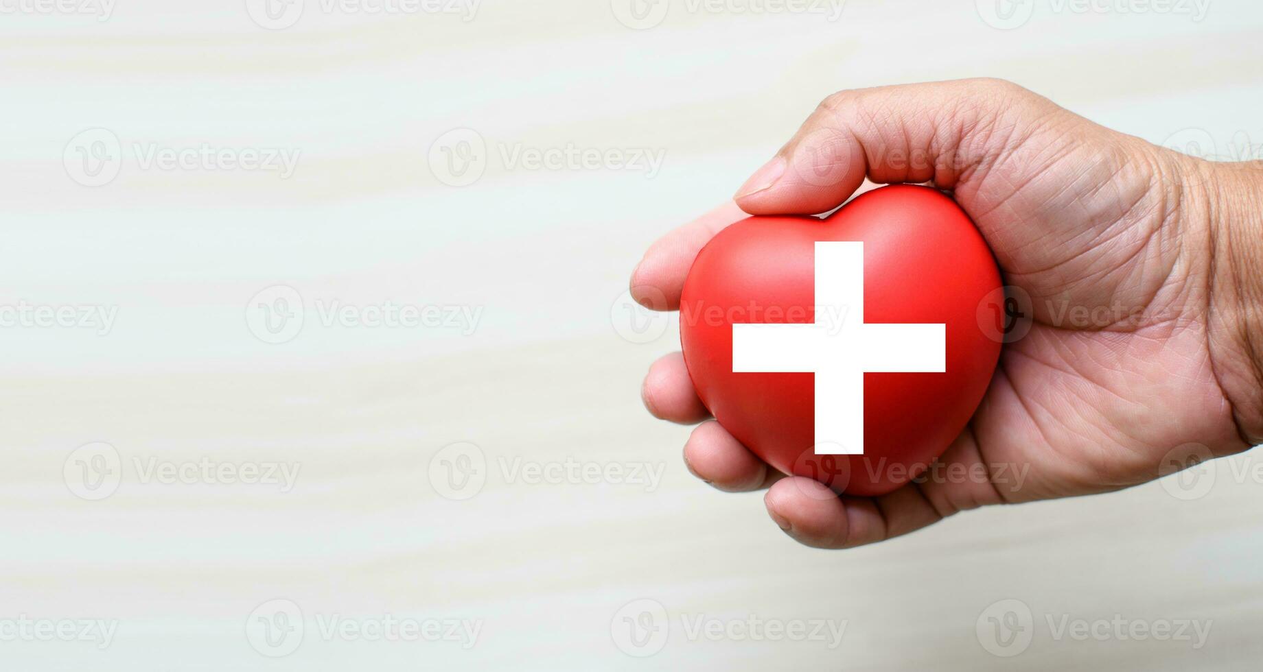 un rojo cruzar es un símbolo de médico salud. mano participación un corazón y en un de madera escritorio. Copiar espacio para el texto y contenido. rojo cruzar RCP, mundo corazón día, mundo salud día, sangre Organo donación foto