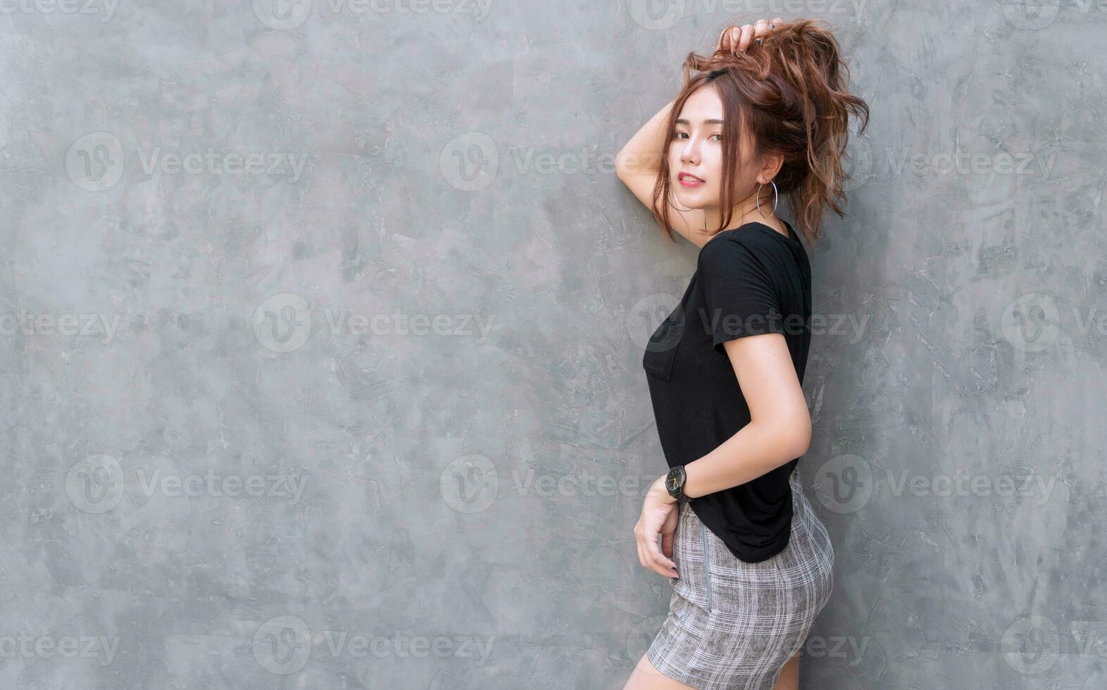 Charming young woman feeling relax holding her thick hair on head over gray wall background. Cheerful girl tying her long brown wavy hair in ponytail looking at something with copy space. photo
