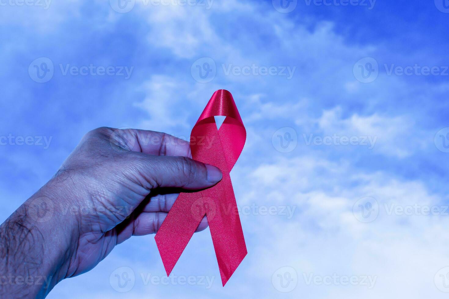 Hand of a man holding red ribbon, HIV awareness concept, world AIDS day, world hypertension day, world cancer day, Take a close-up shot, the background has a blue sky and white cloud photo