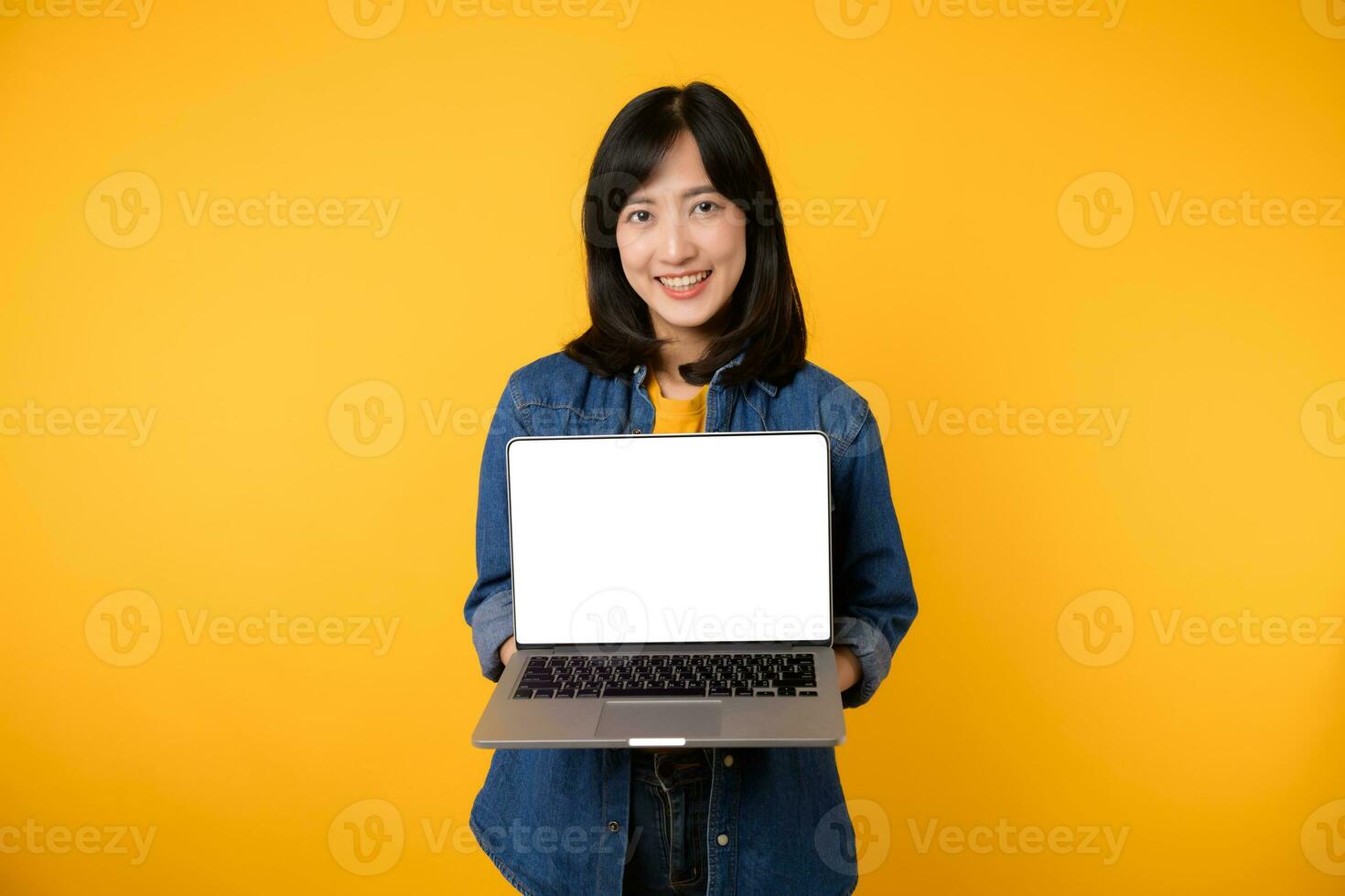 portrait young happy woman wearing yellow t-shirt and denim shirt holding laptop and point finger to screen isolated on yellow studio background. business technology application communication concept. photo