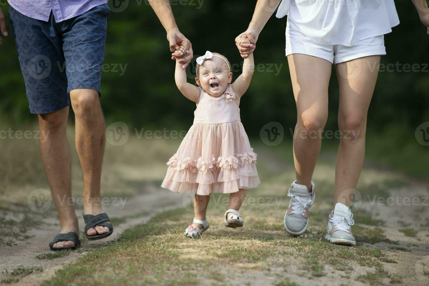 Happy one-year-old girl is being held by dad and mom. Little child walking with parents. A cute baby learns to walk with the help of her parents. photo