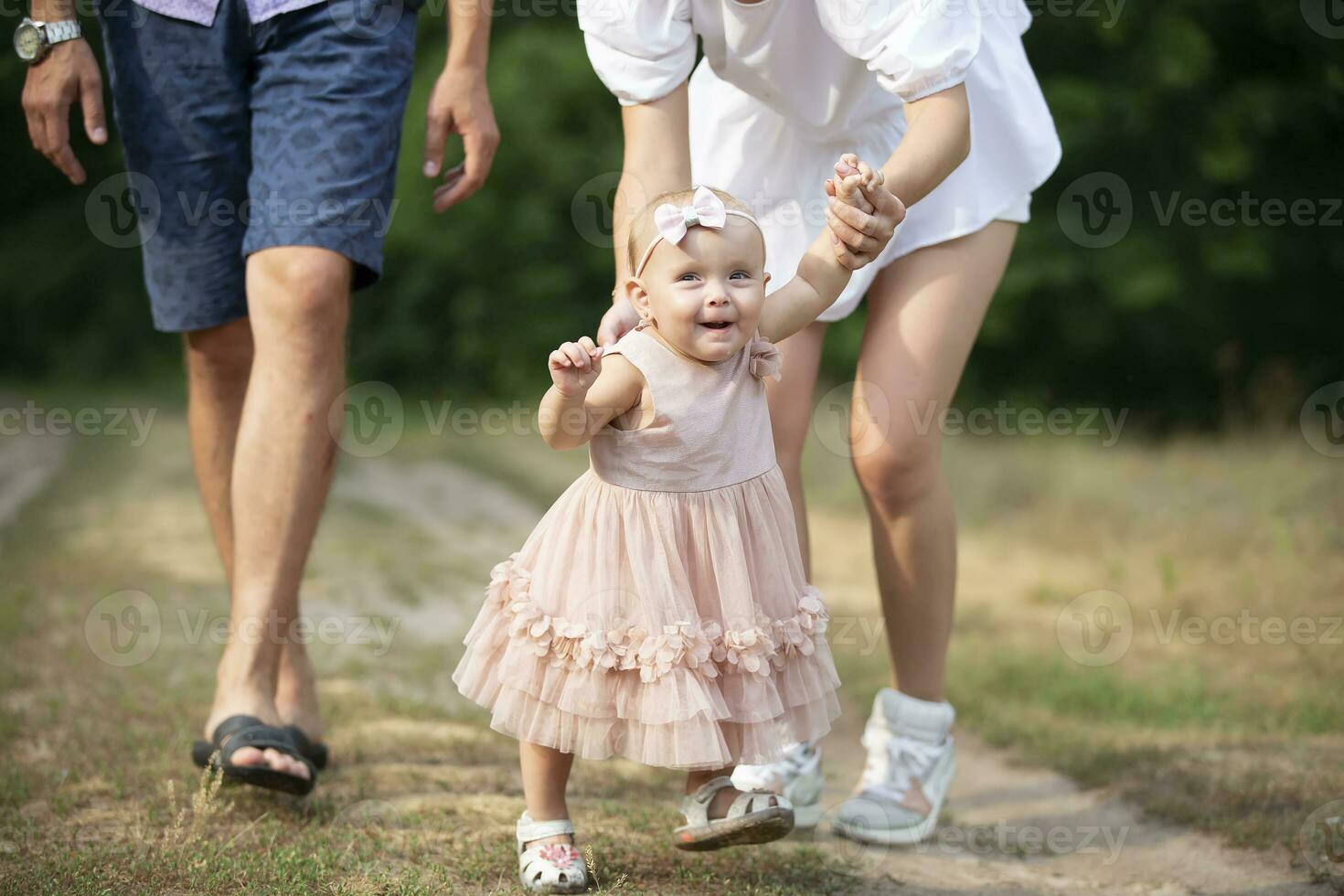 Happy one-year-old girl is being held by dad and mom. Little child walking with parents. A cute baby learns to walk with the help of her parents. photo