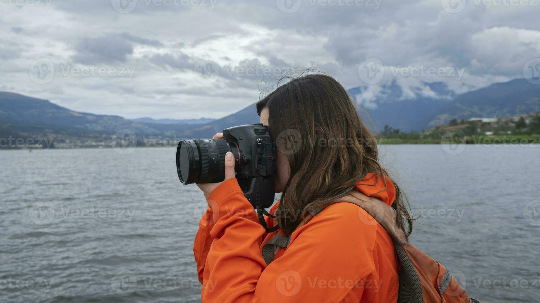 Young Latin American woman dressed in orange jacket with backpack seen from the side taking photos next to a lake surrounded by mountains