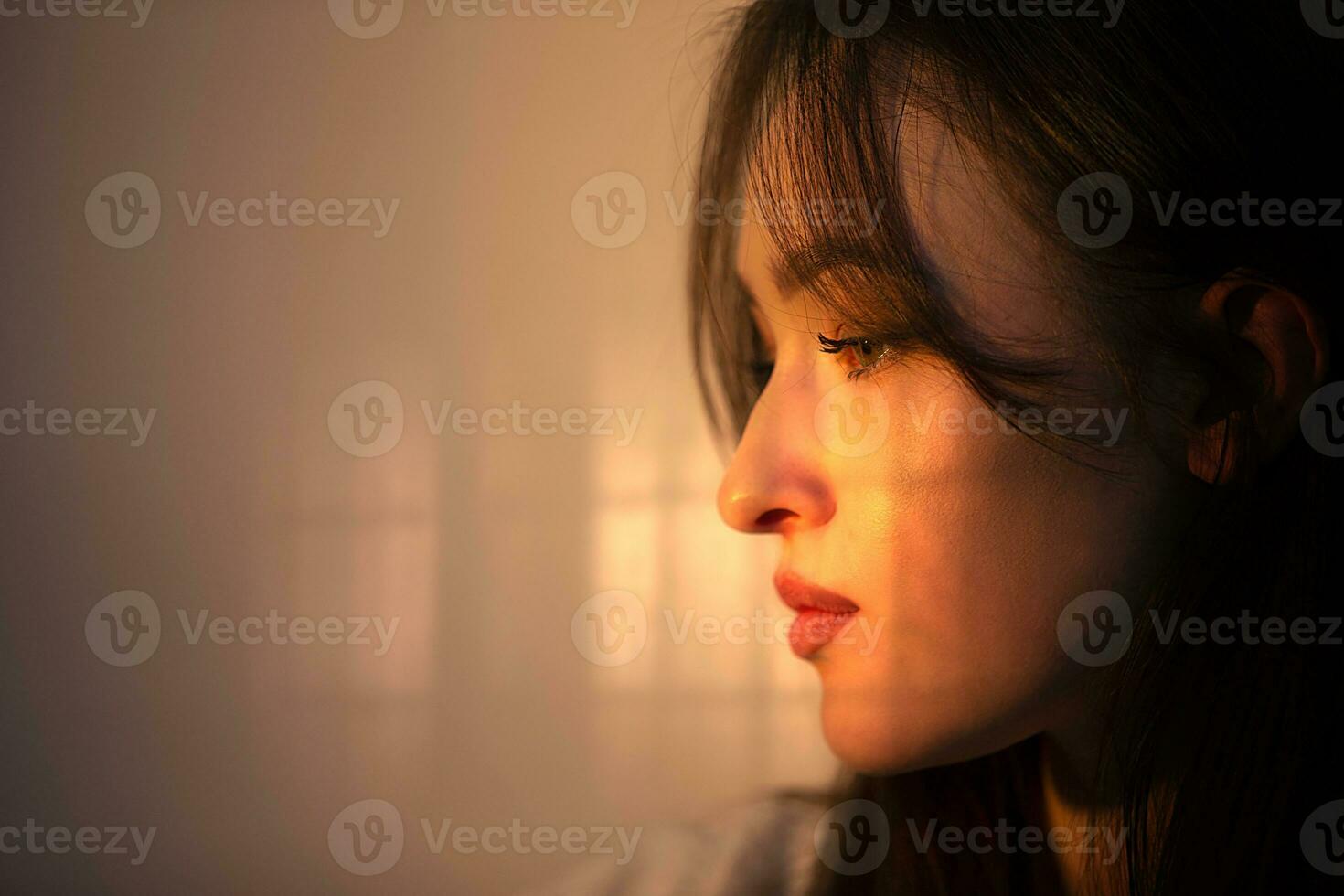 The face of a pensive girl in profile is illuminated by warm light from the window. photo