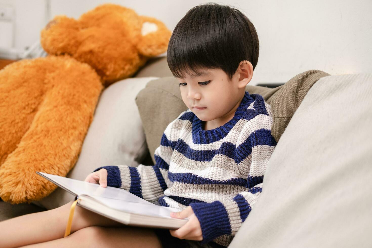 asiático chico leyendo un libro en el sofá aprendizaje fuera de el salón de clases foto