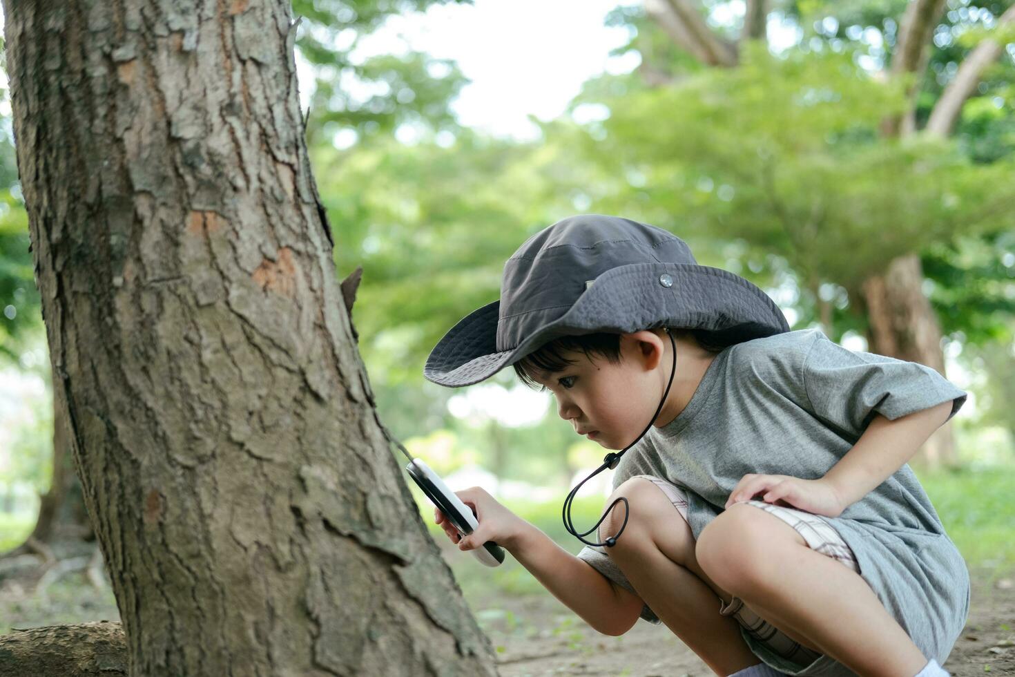 Asian boy wearing a hat in a forest exploration suit Use a magnifying glass to survey the tree area. photo