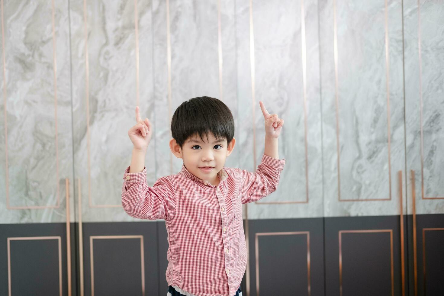 An Asian boy stands and smiles, raising both hands and pointing upwards. photo