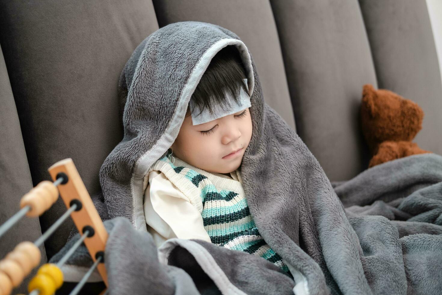 asiático chico acostado enfermo, él tenido un fiebre reduciendo parche en su frente y cubierto él mismo con un cobija porque de el frío. foto