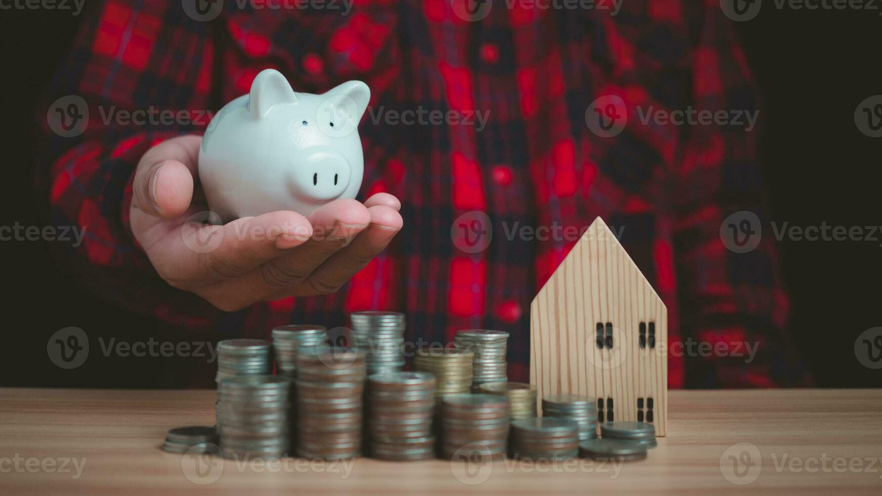 Human holds a piggy bank and there are coins and model wooden houses lined up on the wooden floor. Concepts of finance, savings, investment, and setting goals for fixed deposits with banks. photo