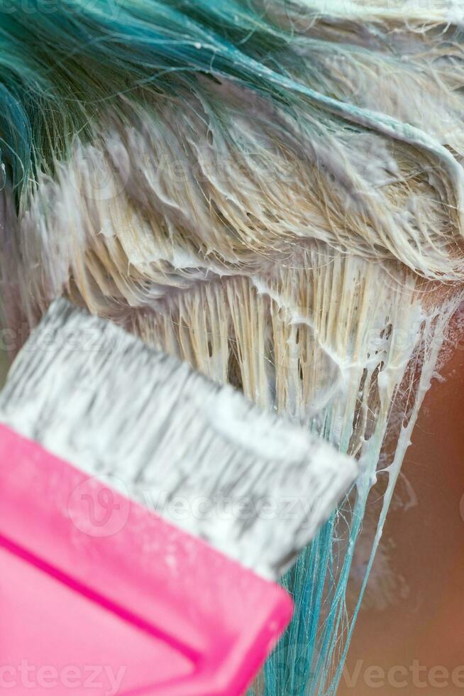 Hairdresser using pink brush while applying paint to female customer with emerald hair color during process of bleaching hair roots photo