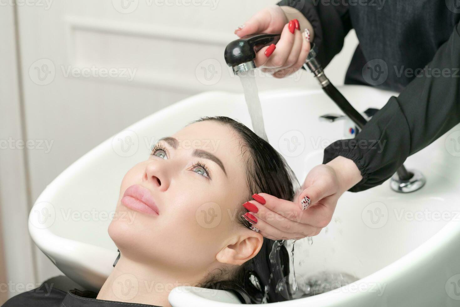 Hairstylist's hands wash long hair of brunette woman with shampoo in special sink for shampooing photo