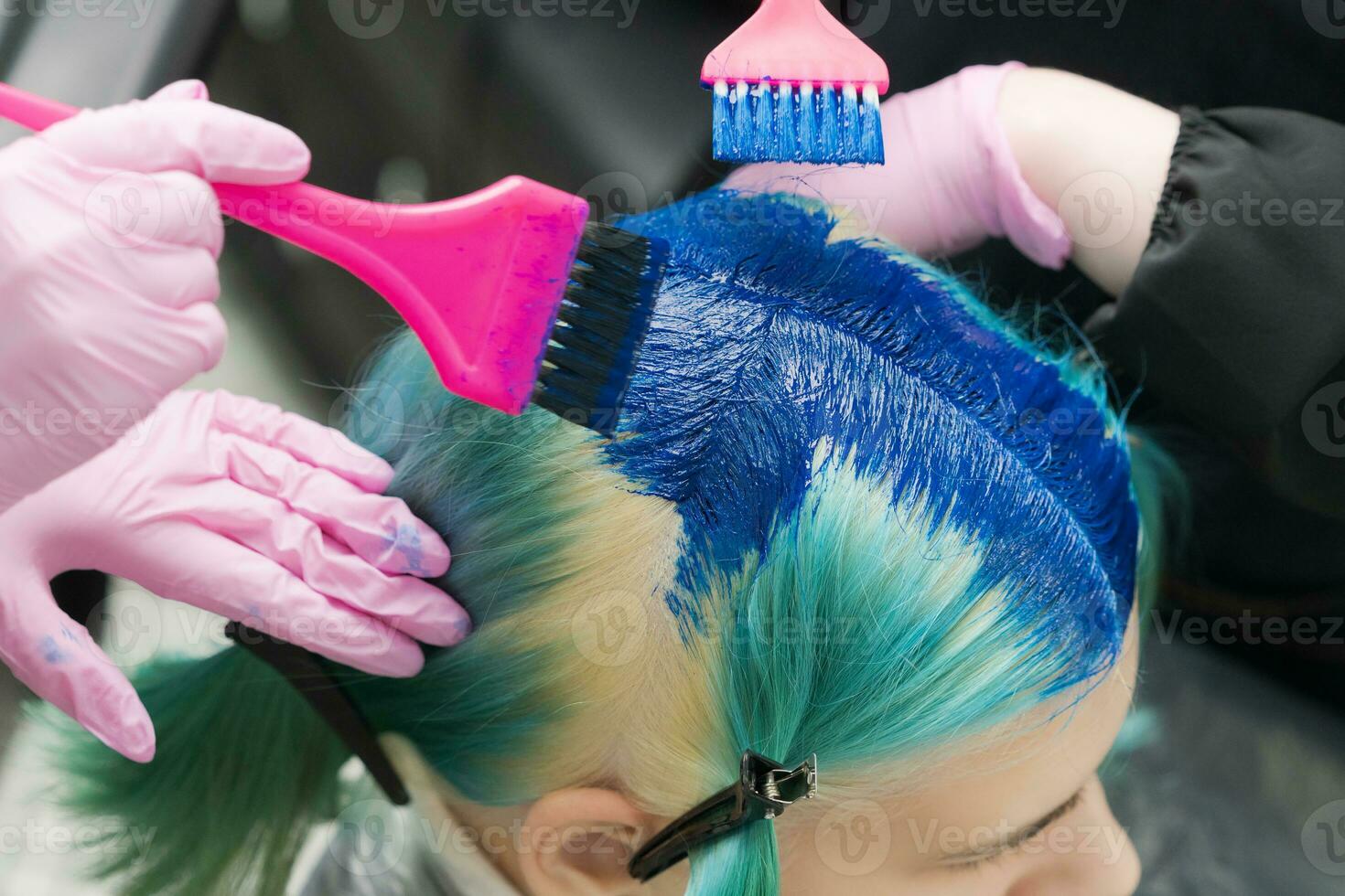 Two hairdressers using pink brush while applying blue paint to female with emerald hair color during process of dyeing hair in unique lapis lazuli color photo