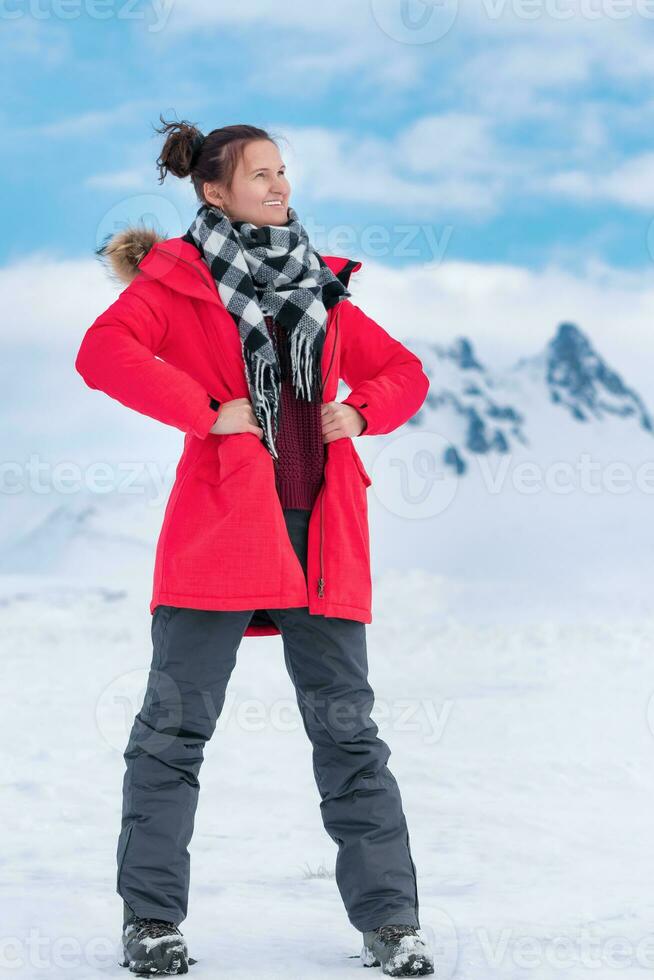 Woman tourist dressed in red winter jacket, sports pants, trekking boots stands in winter mountains photo