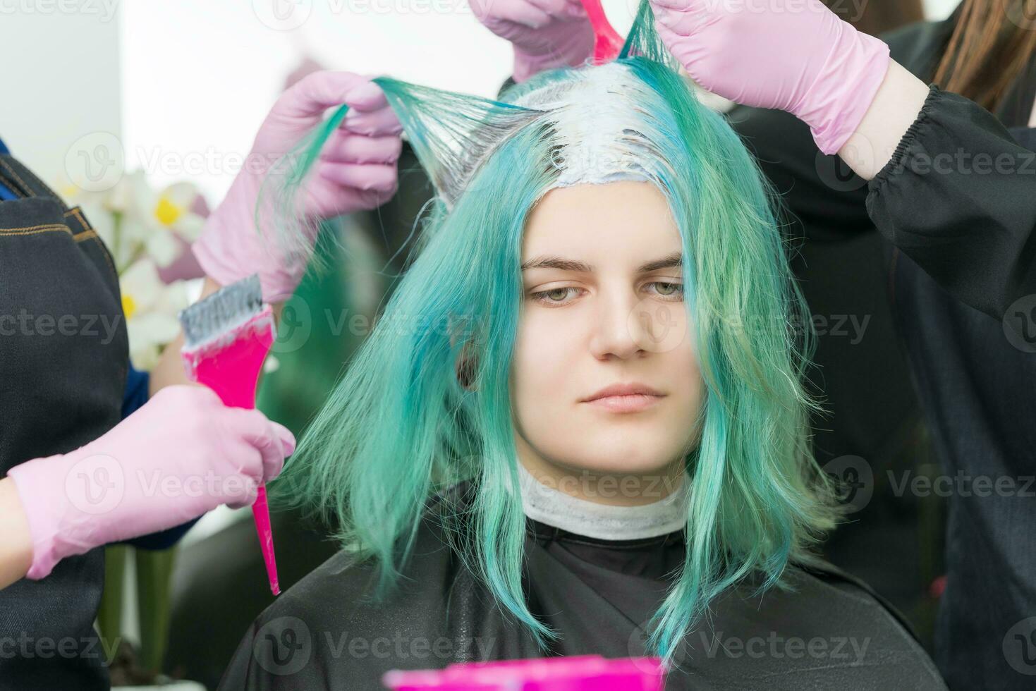 Two hairdressers use pink brush when applying paint to hair during process of bleaching hair roots. Young adult customer with long emerald hair color in beauty salon photo