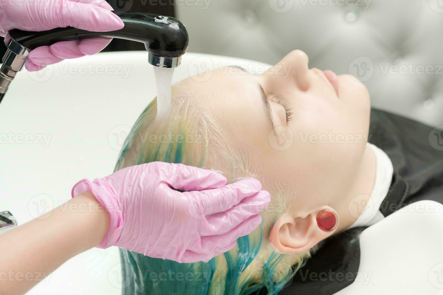 Hairdresser washes client's head with green hair color. Hairstylist washing hair with shampoo of woman in sink with shower in beauty salon photo