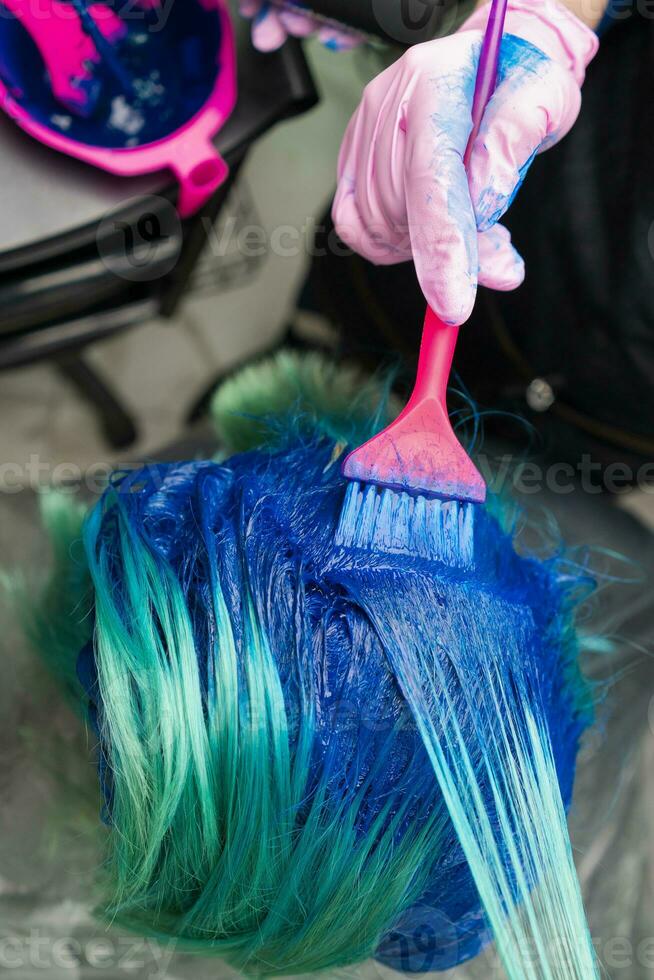 Hairdresser in glove using pink brush while applying blue paint to customer during process of dyeing hair in stylish color photo