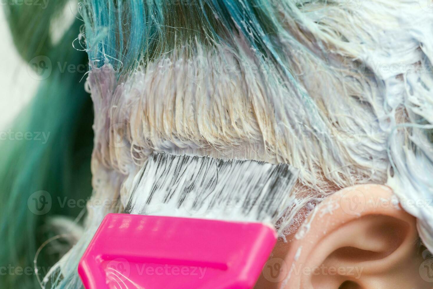 Hairdresser using pink brush while applying paint to female customer with emerald hair color during process of bleaching hair roots photo