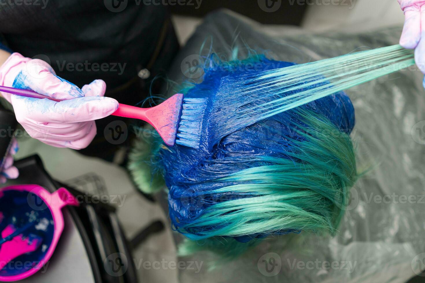 peluquero en protector guante utilizando rosado cepillo mientras aplicando azul pintar a cliente durante proceso de tintura pelo en único color foto