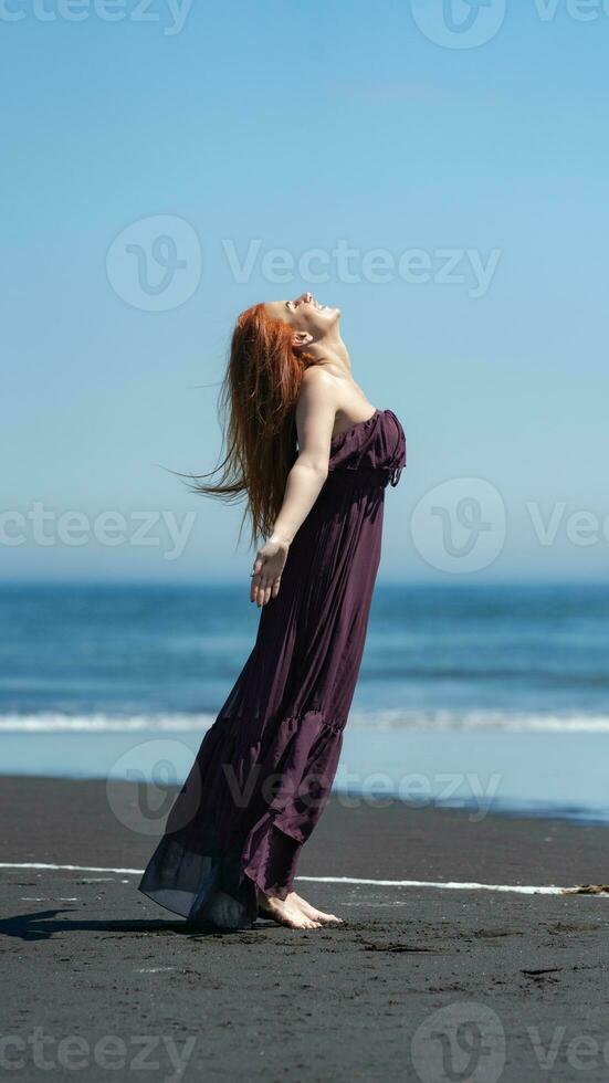 Happy woman in long dress stands on beach with head looking upward, eyes closed on background of sea photo