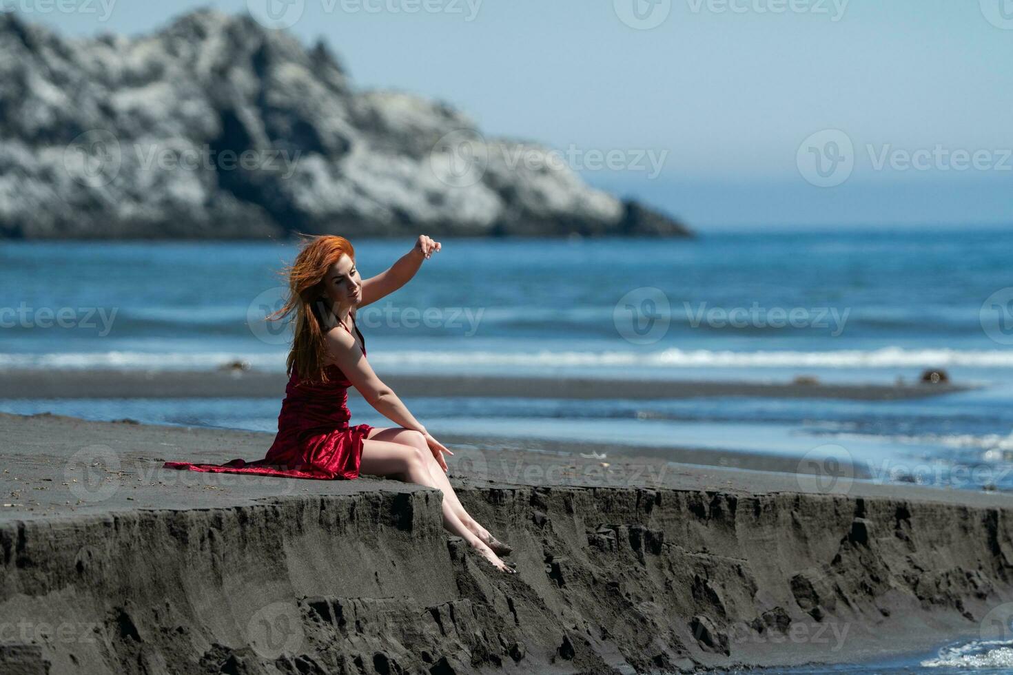 Woman in red long dress with raised hem sitting on sandy beach with bare legs dangling over cliff photo