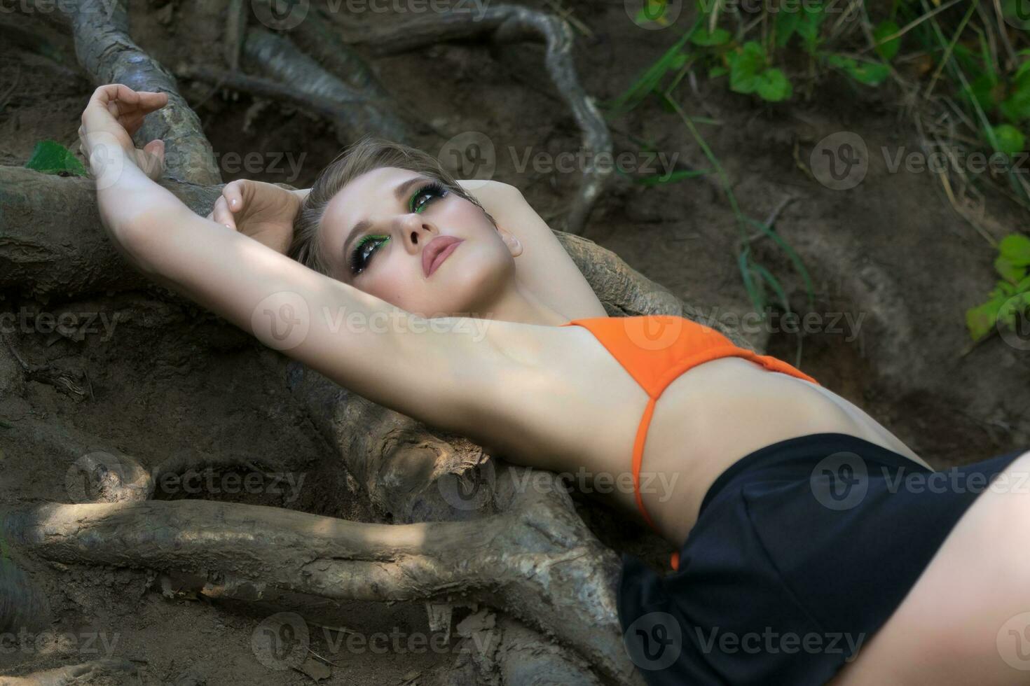 sensual mujer acostado abajo en suelo en árbol raíces en bosque. Delgado hembra en bikini parte superior y mini falda foto