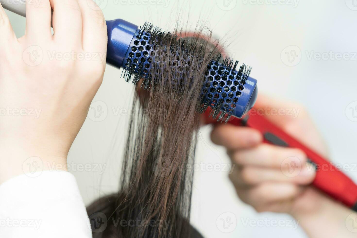 Hands of hairdresser dries brunette hair using blue hairbrush, red hairdryer in beauty salon photo