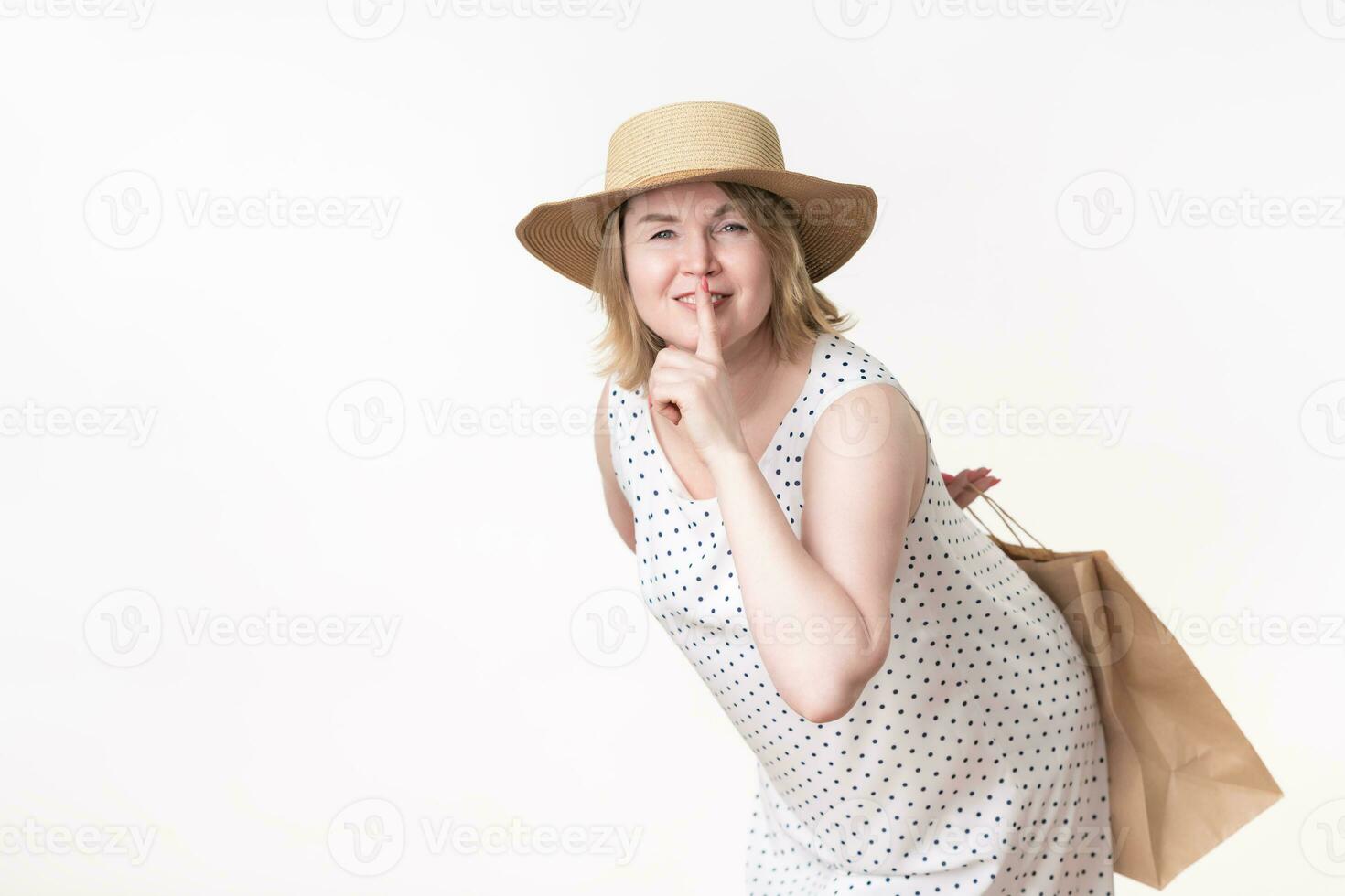 Playful woman shopper put her index finger to lips and holding craft paper shopping bag behind back photo