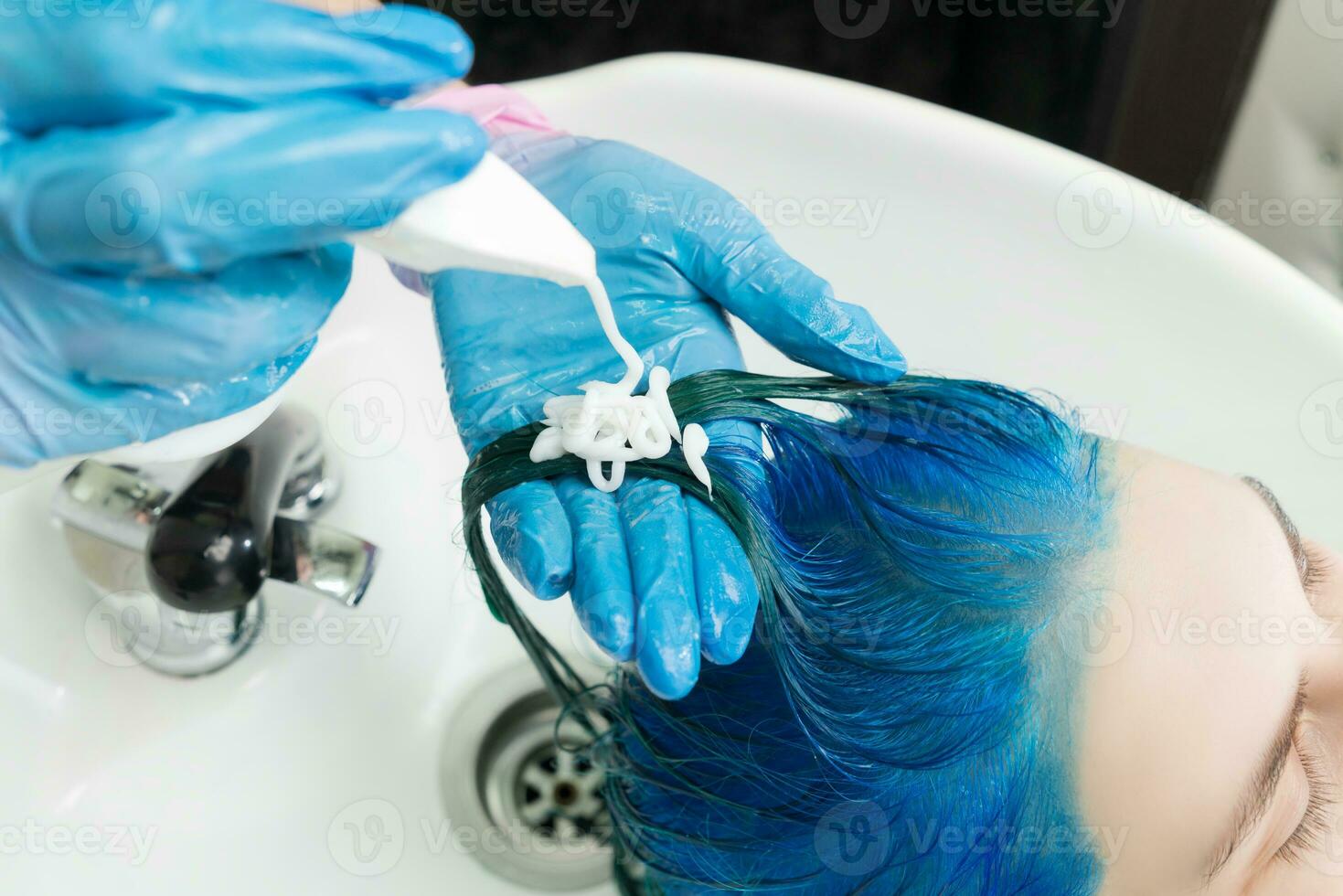 peluquero apretones champú desde tubo sobre clientela azul pelo mientras Lavado pelo en lavabo y ducha a belleza salón foto