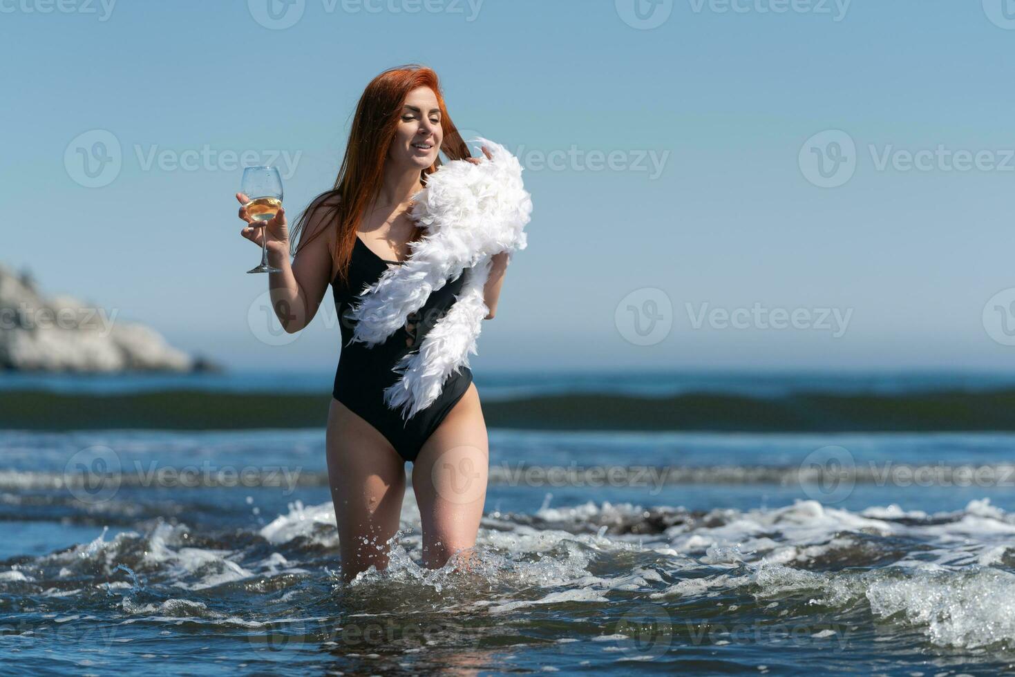 Sensuality woman in swimsuit walks knee deep in waves of ocean, holding wine glass and boa in hands photo