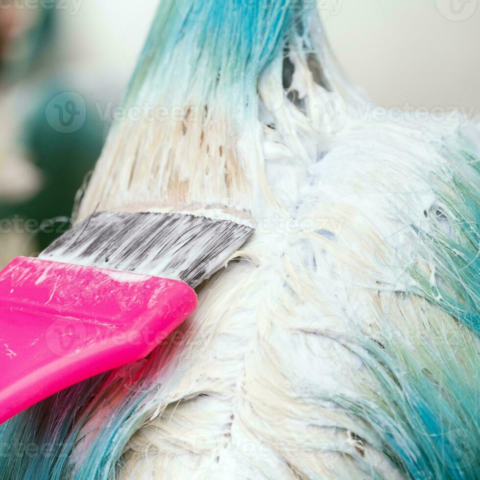 Stylist using pink brush while applying paint to female customer with emerald hair color during process of bleaching hair roots photo